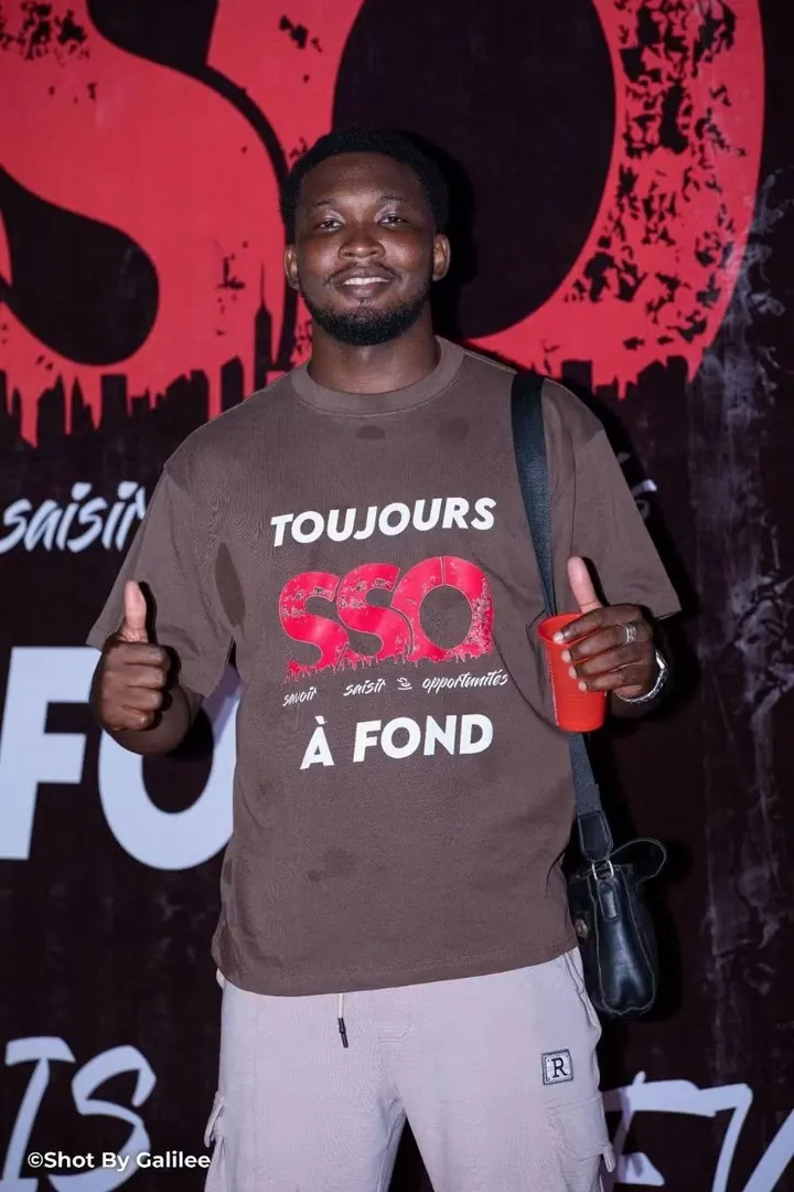 Dyjor, membre de la team SSO, devant le mur SSO en t-shirt marron, pouces levés
